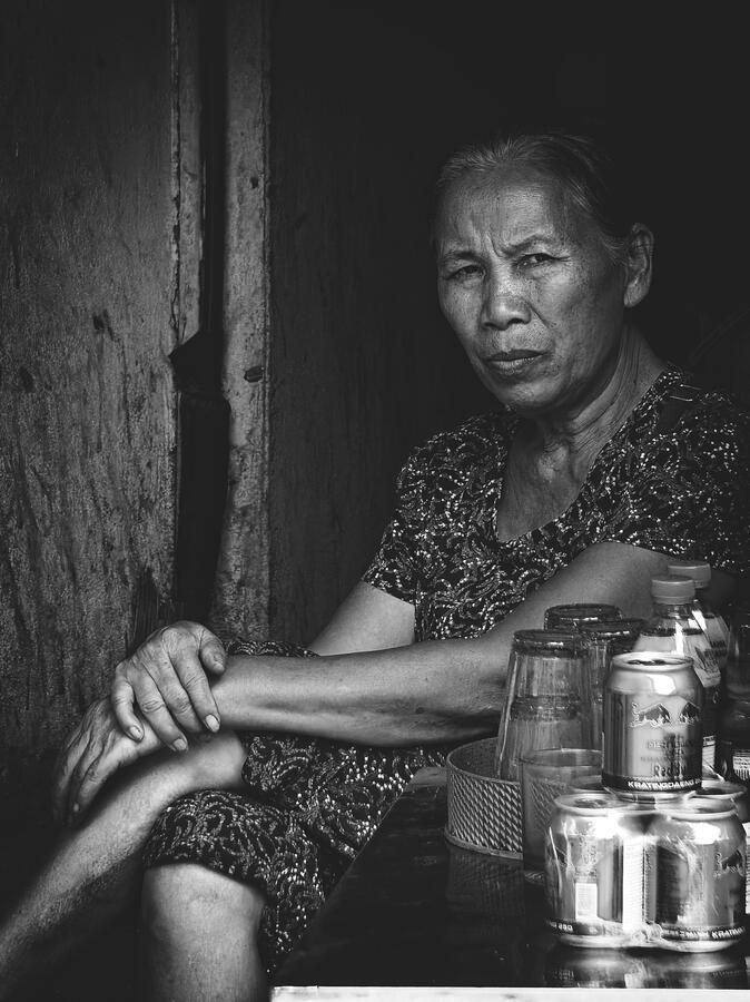 A Moment of Stillness A woman sits quietly in a shaded doorway, her gaze meeting the camera. Captured in black and white, the portrait reveals the textures, expressions and quiet dignity of everyday life in Vietnam.