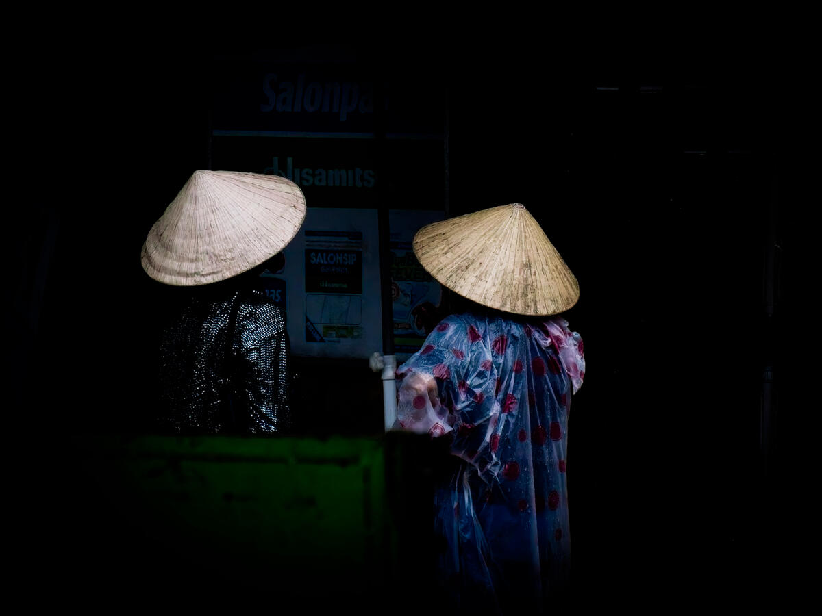 Two Hats Two figures wearing traditional Vietnamese conical hats emerge from the darkness, their clothing and silhouettes illuminated by the available light. A quiet, unscripted moment of everyday life in Vietnam.