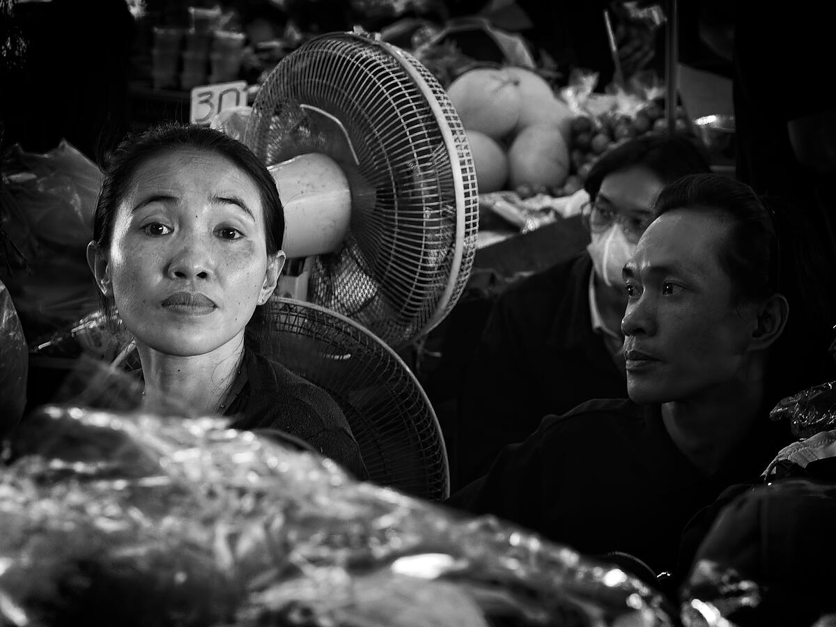 Three market workers during a quiet moment in Vientiane, Laos. Black and white photograph of three market workers surrounded by produce and electric fans in Vientiane, Laos.