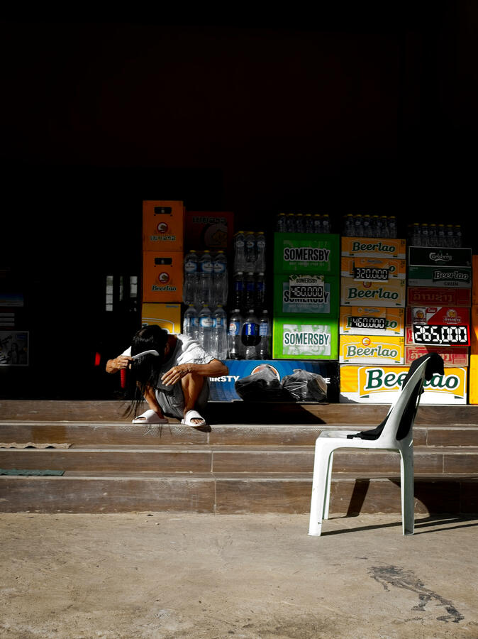 A woman dries her hair in the afternoon sun outside a shop in Vientiane, Laos. Woman crouching in strong sunlight outside a shop surrounded by Beerlao crates and an empty plastic chair in Vientiane, Laos.