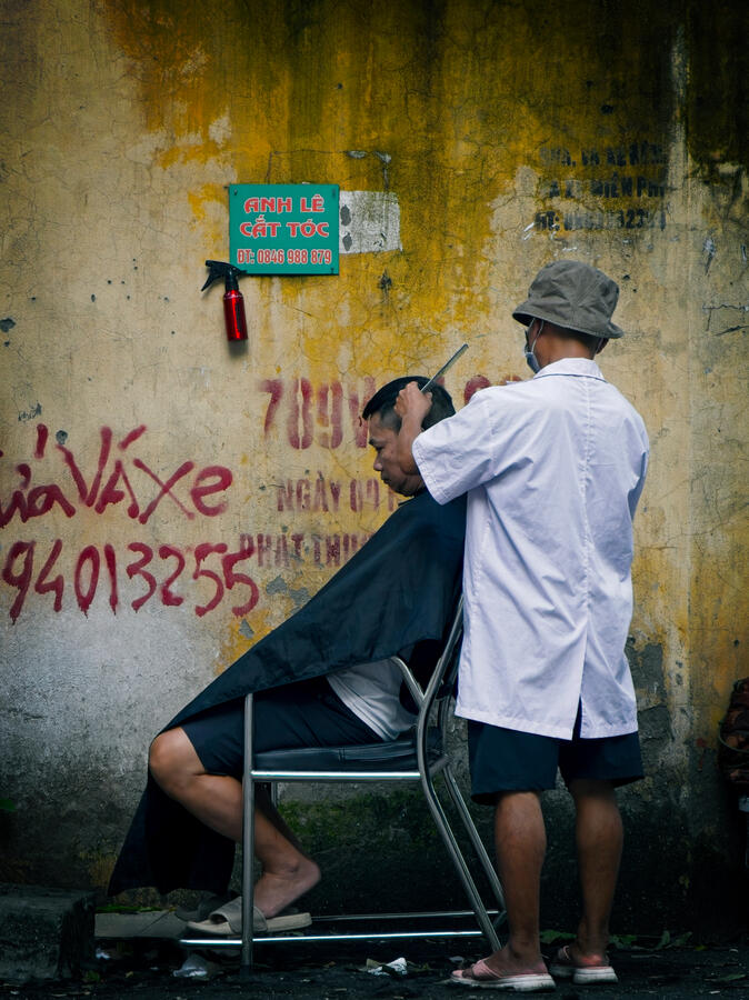 A roadside haircut against the weathered walls of Hanoi, Vietnam. Street barber cutting a man's hair beside a weathered yellow wall in Hanoi, Vietnam.
