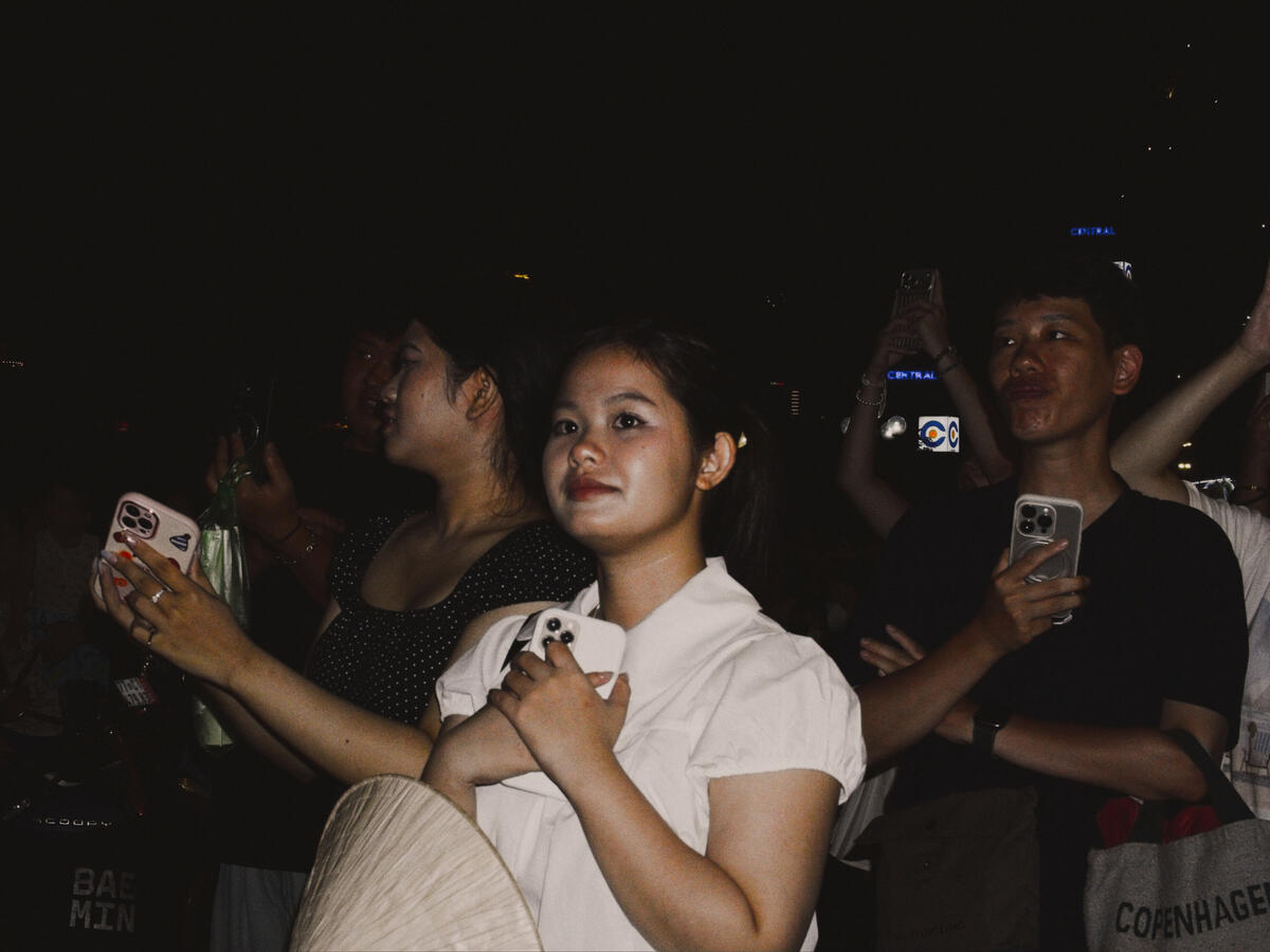 In the Moment A crowd gathers beneath the night sky in Da Nang, Vietnam, their faces illuminated by the camera's flash. Surrounded by people capturing the occasion on their phones, a fleeting moment of anticipation becomes a portrait of modern street life.