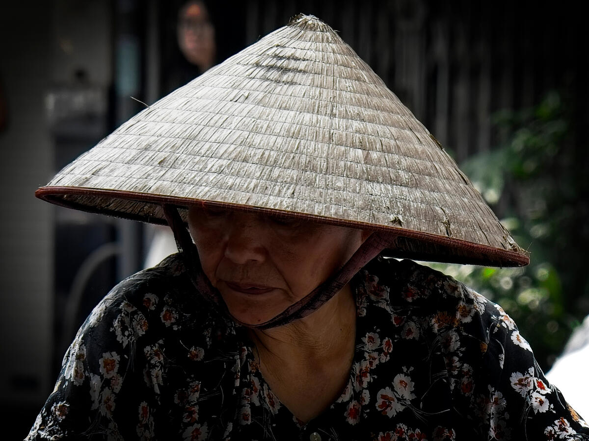 A street vendor beneath a traditional conical hat in Hanoi, Vietnam. Close portrait of an older Vietnamese woman wearing a traditional conical hat in Hanoi, Vietnam.